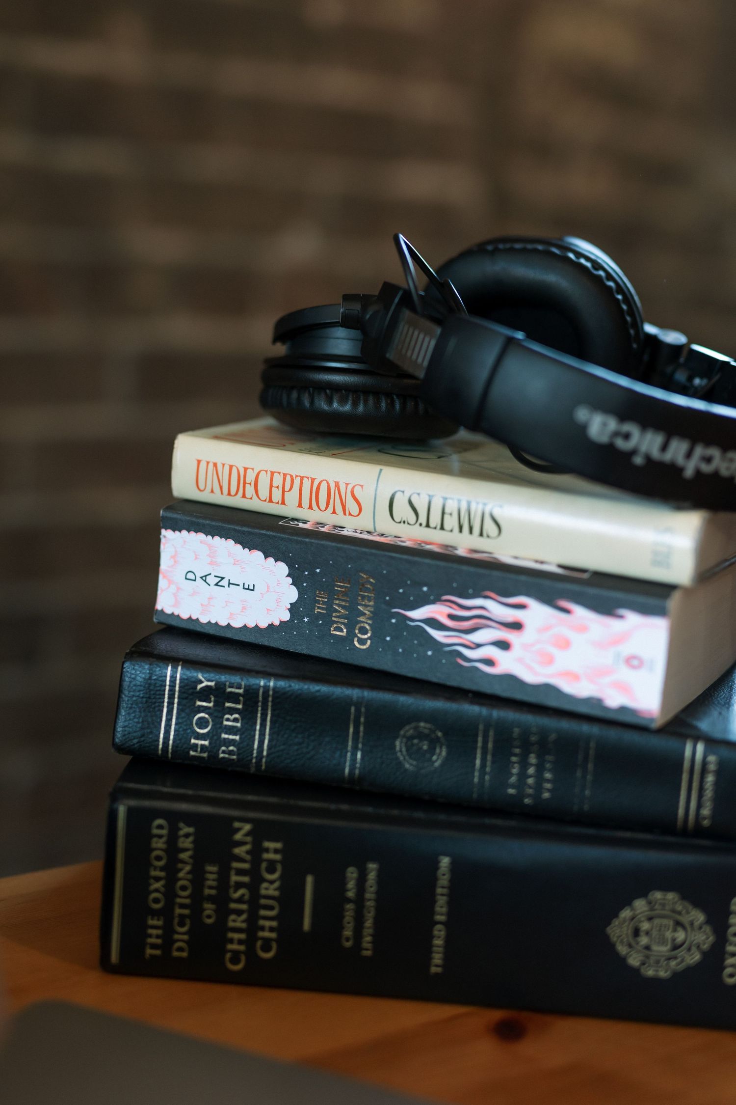 Stack of books including Holy Bible and CS Lewis title with black Audio-Technica headphones on wooden surface.