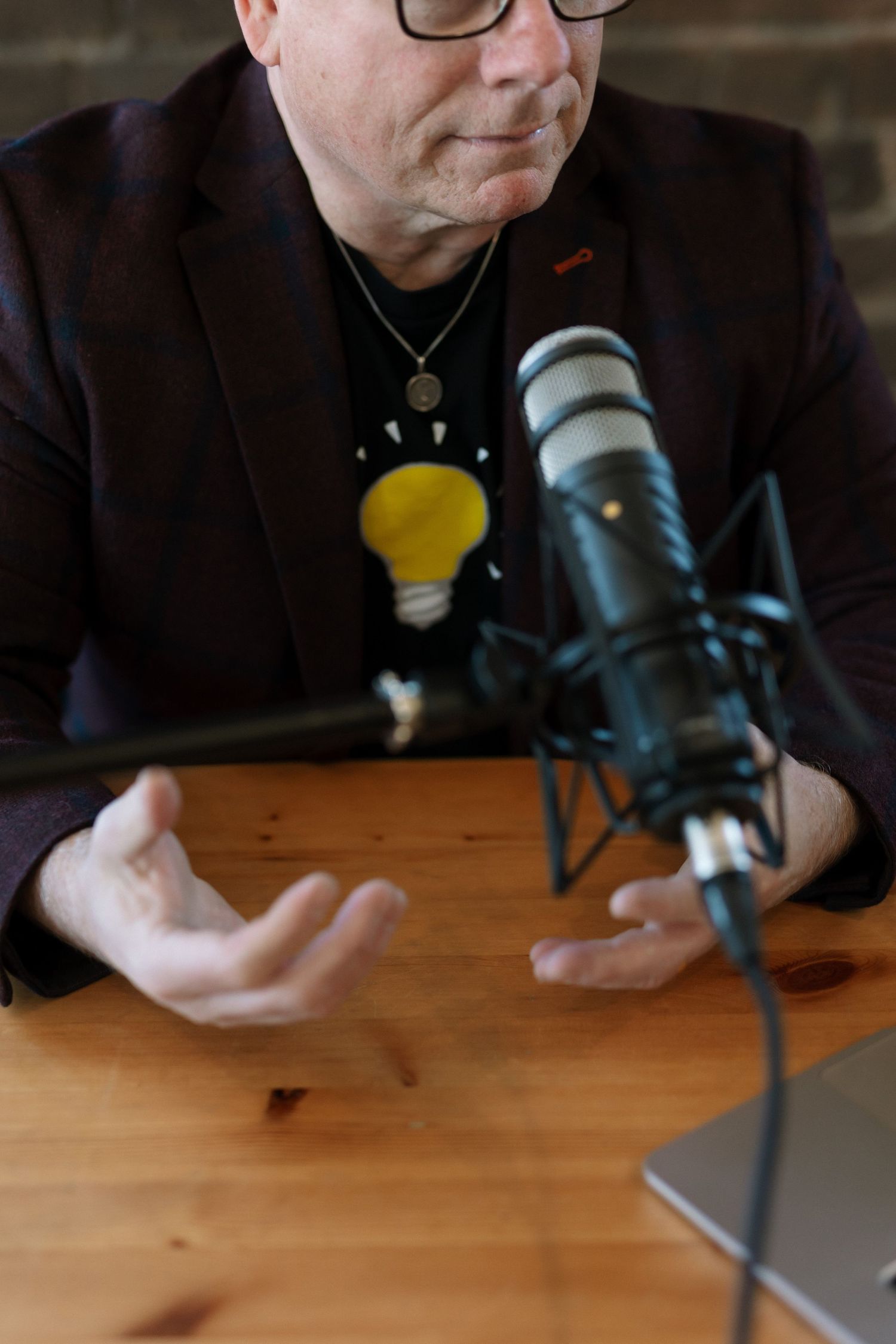 Man in blazer gestures while speaking into studio microphone at wooden desk, wearing lightbulb graphic tee.