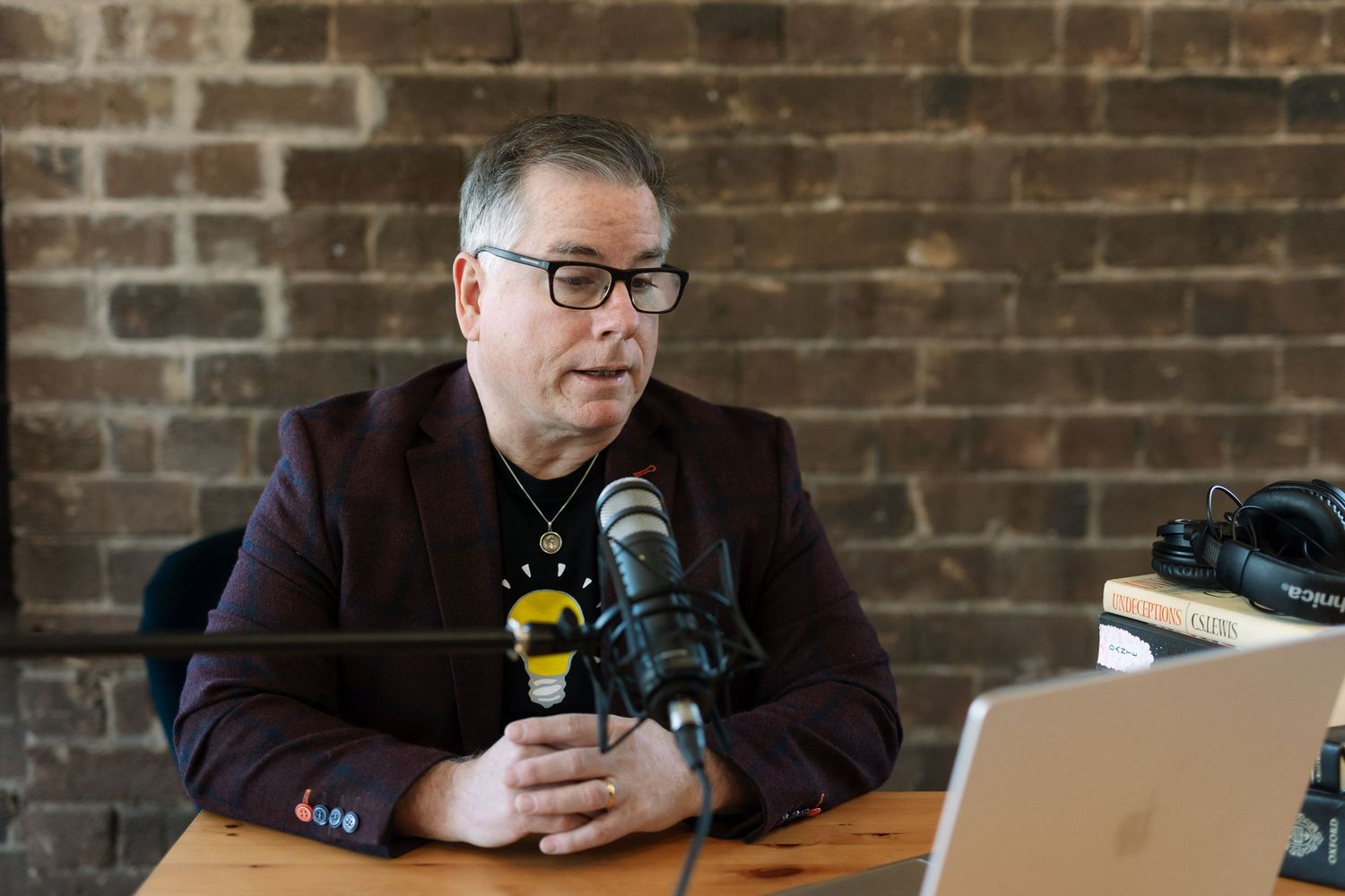 Man in blazer speaking into podcast microphone at wooden desk with laptop, brick wall background.