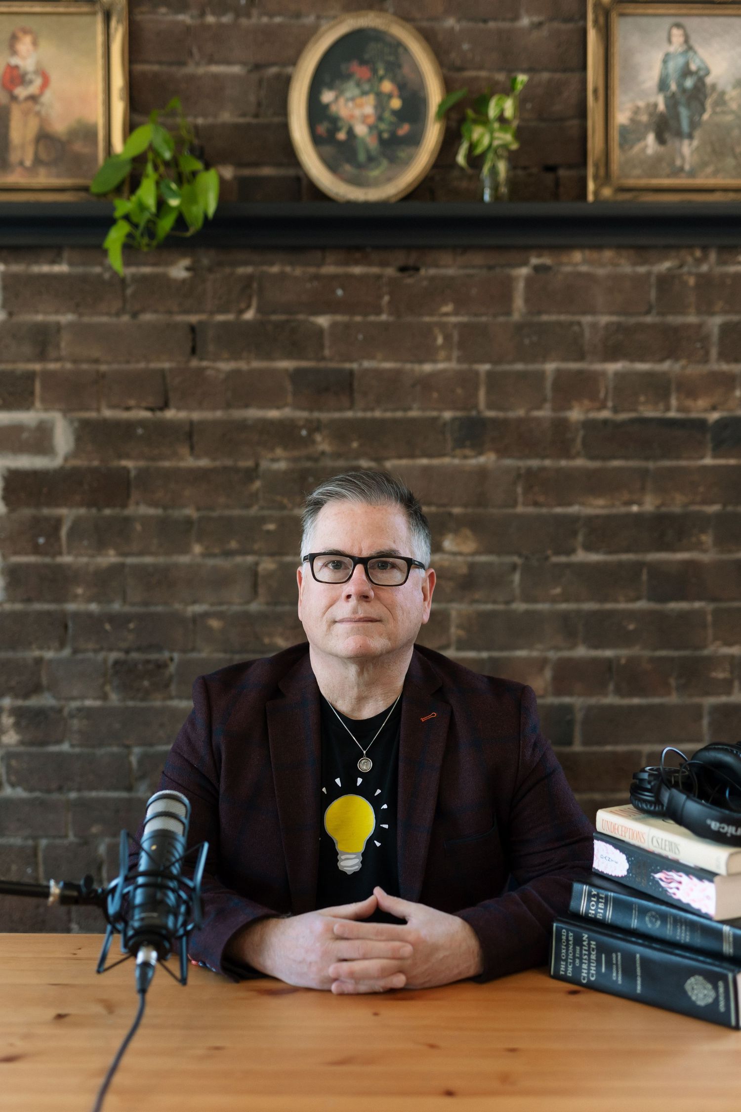 Man in dark blazer sits at wooden table with microphone and books, brick wall and vintage paintings behind him.