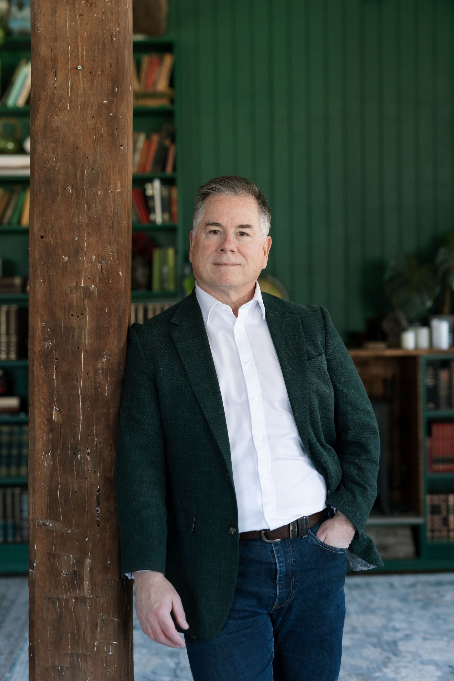 Middle-aged man in green blazer and jeans leaning against wooden post in front of green bookshelf.