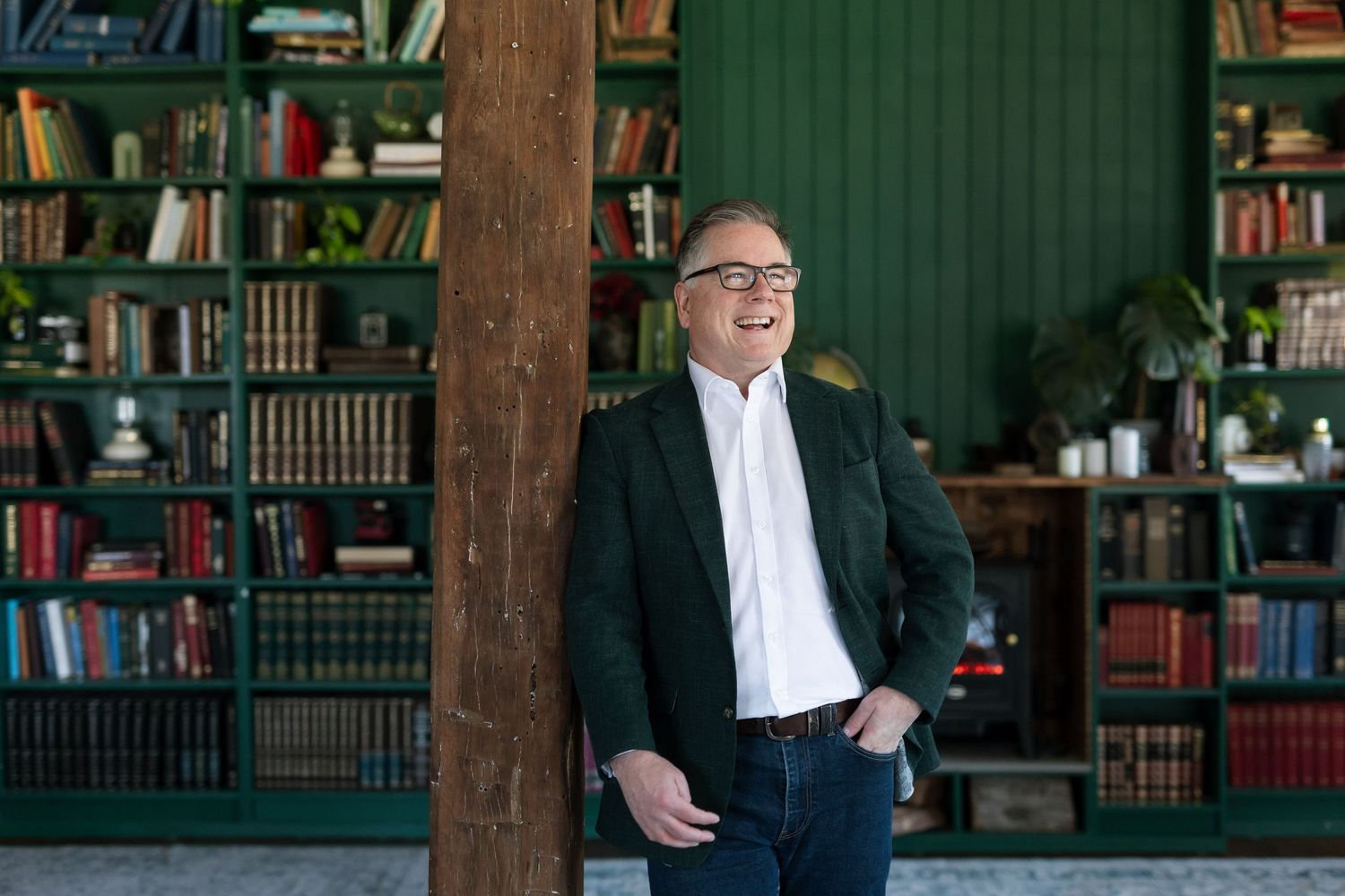 Man in green blazer smiling in a cozy library with green bookshelves filled with books.