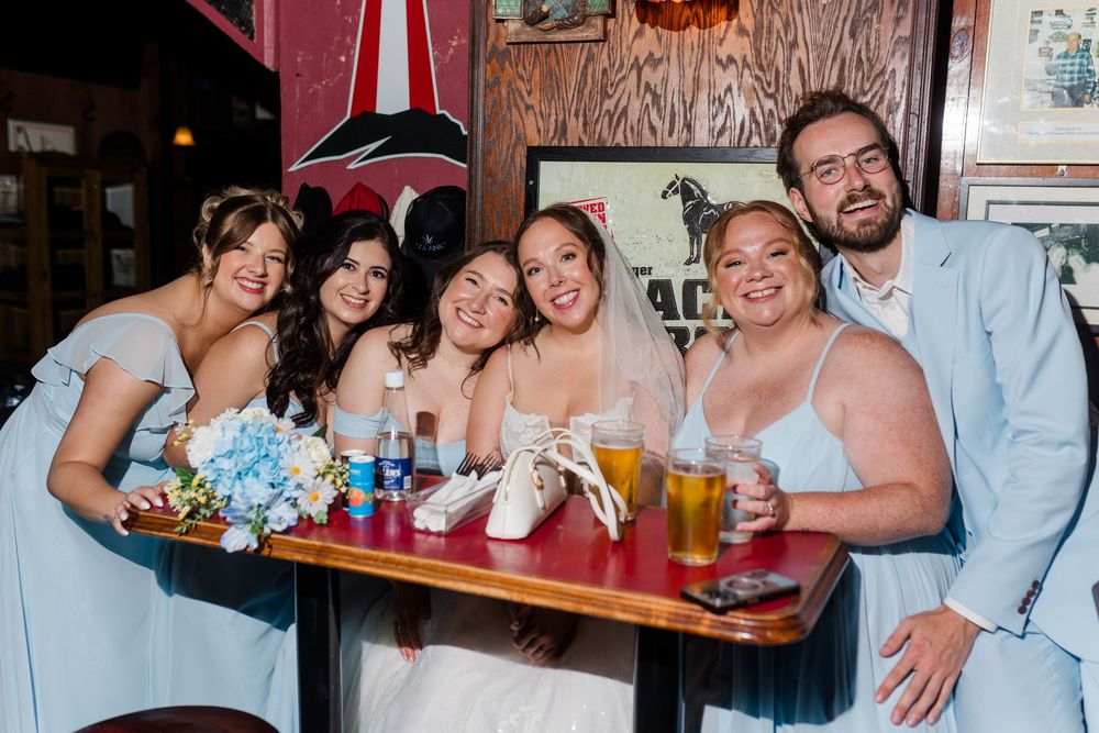 Bride in white gown poses with bridesmaids in light blue dresses and a groomsman at a bar table.