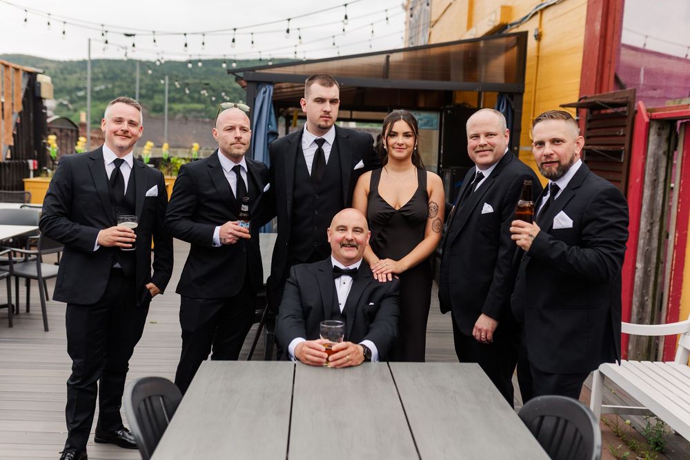 A group of groomsmen in black suits pose with drinks on an outdoor patio at a wedding celebration.