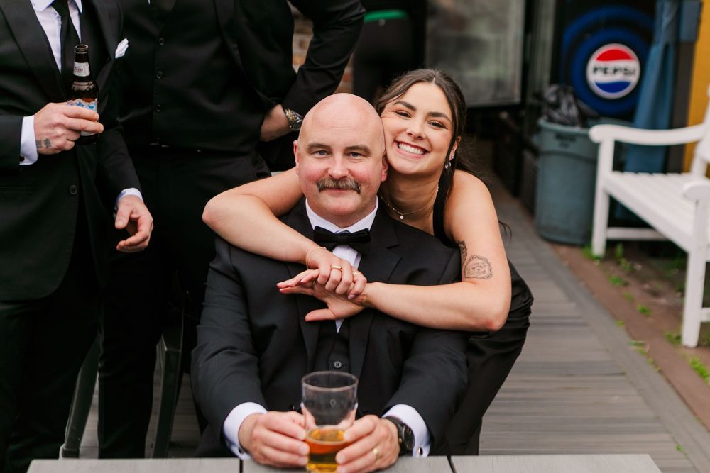 A smiling woman hugs a bald man in a tuxedo at a wedding reception, both posing happily at an outdoor table.