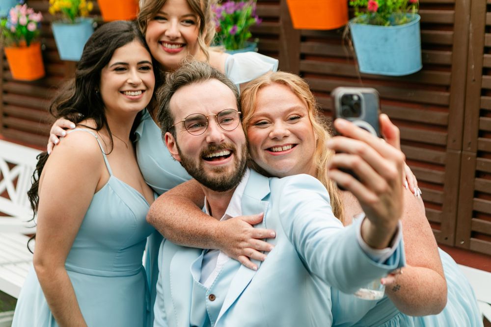 Group of four friends in light blue outfits taking a joyful selfie at an outdoor wedding celebration.
