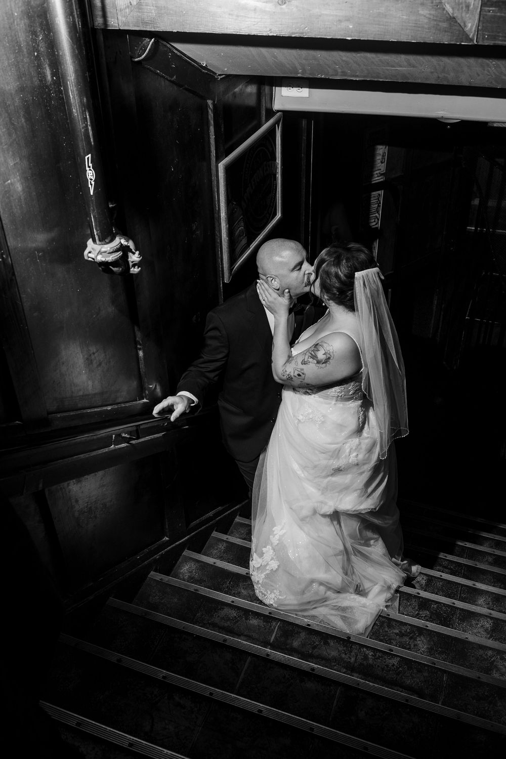 Black and white photo of a bride and groom sharing a kiss on a staircase, bride in lace gown with veil.