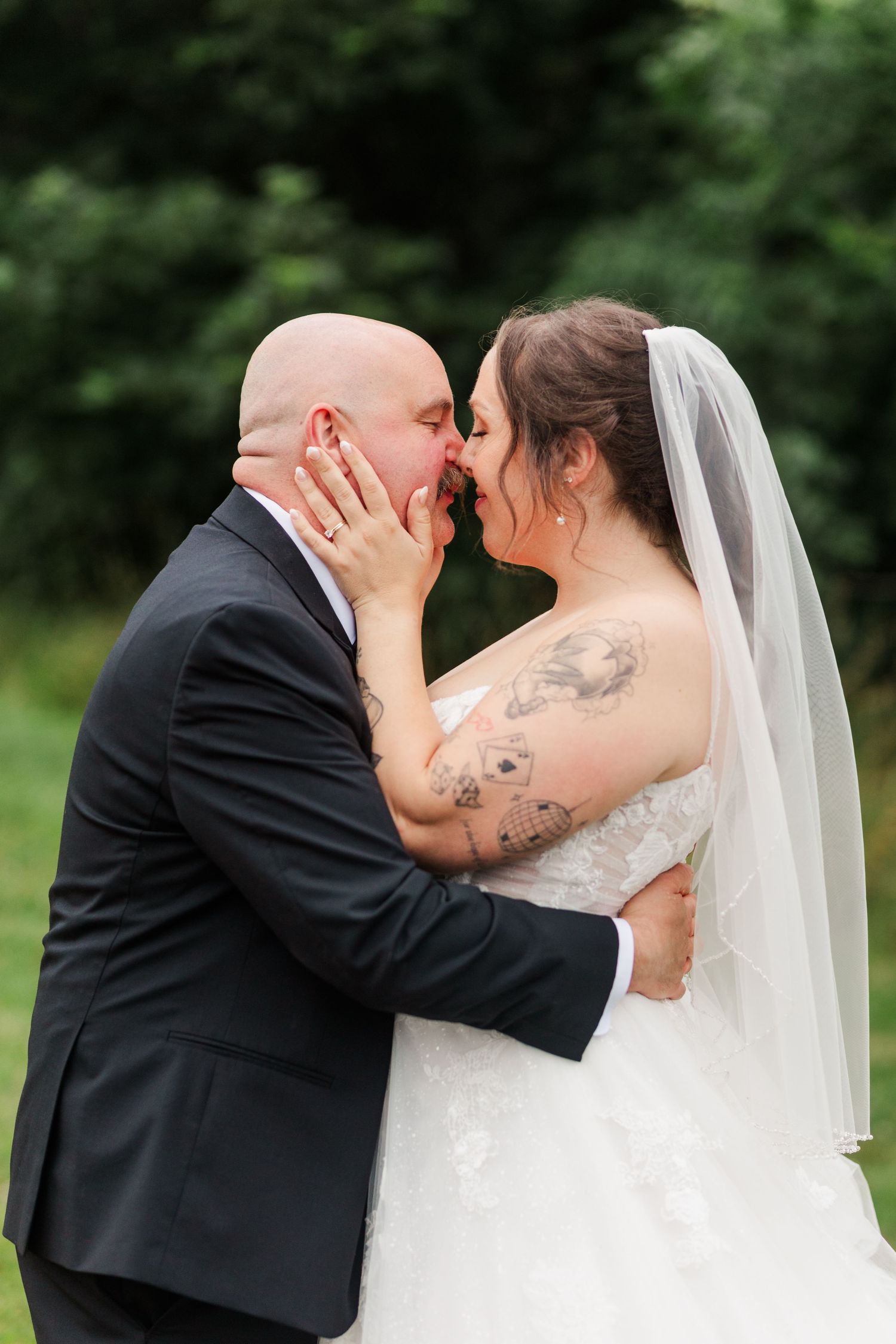 A bride and groom share a romantic kiss outdoors, with the bride in a lace gown and veil, showcasing her tattoos.