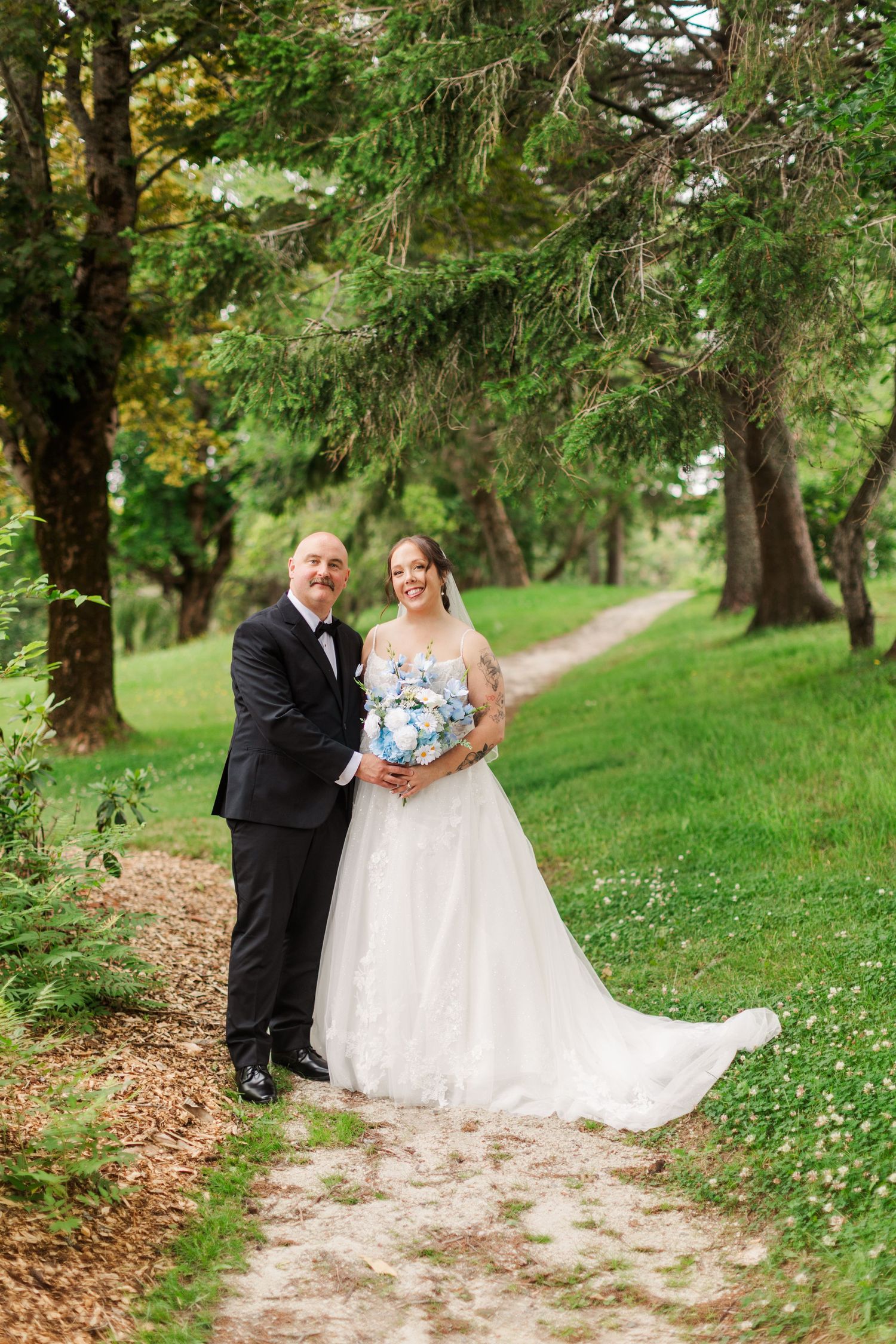 Bride and groom smiling on a tree-lined path, bride holding blue and white bouquet in elegant white gown.