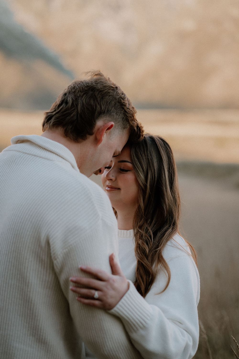 Couple in matching white sweaters embracing tenderly outdoors with mountain landscape in background.