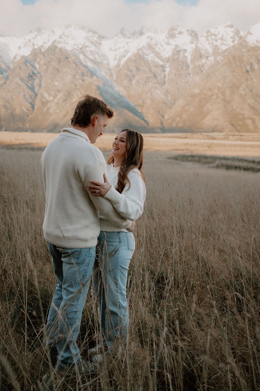 Couple embracing in golden field with snow-capped mountains, both wearing white sweaters and jeans.