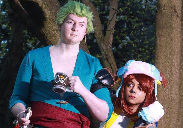 Two cosplayers in anime costumes pose outdoors, one in teal holding a goblet, another in colorful outfit with red hair.