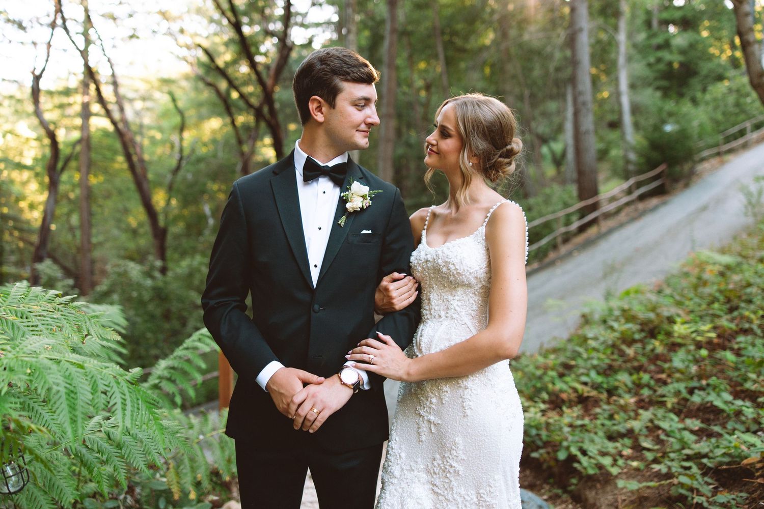 Bride and groom pose together outdoors in lush forest setting, groom in black tuxedo, bride in lace wedding gown.