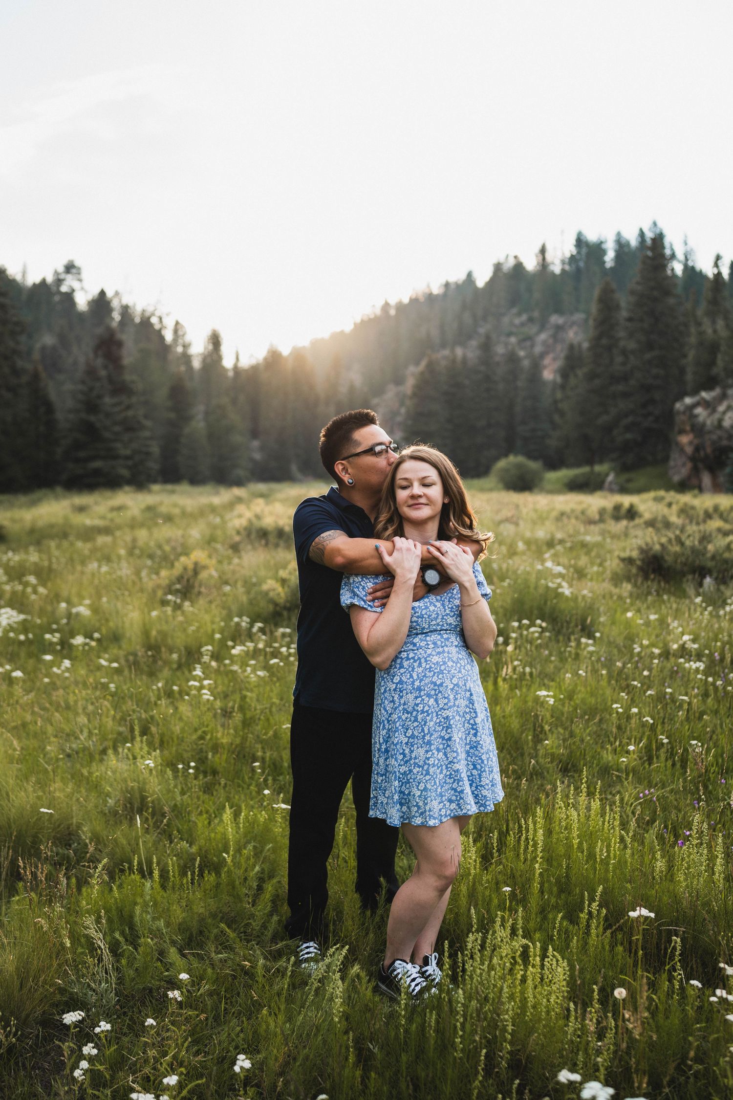 Couple embracing in a sunlit mountain meadow with wildflowers, man kissing woman's cheek, evergreen trees in background.