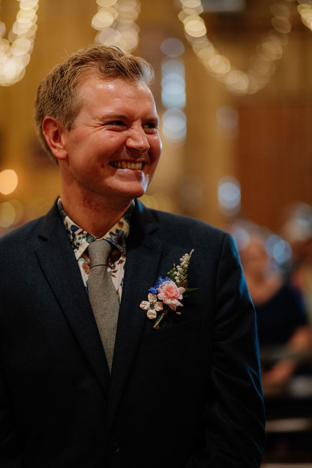 Smiling groom in navy suit with floral boutonniere and patterned shirt at wedding ceremony.