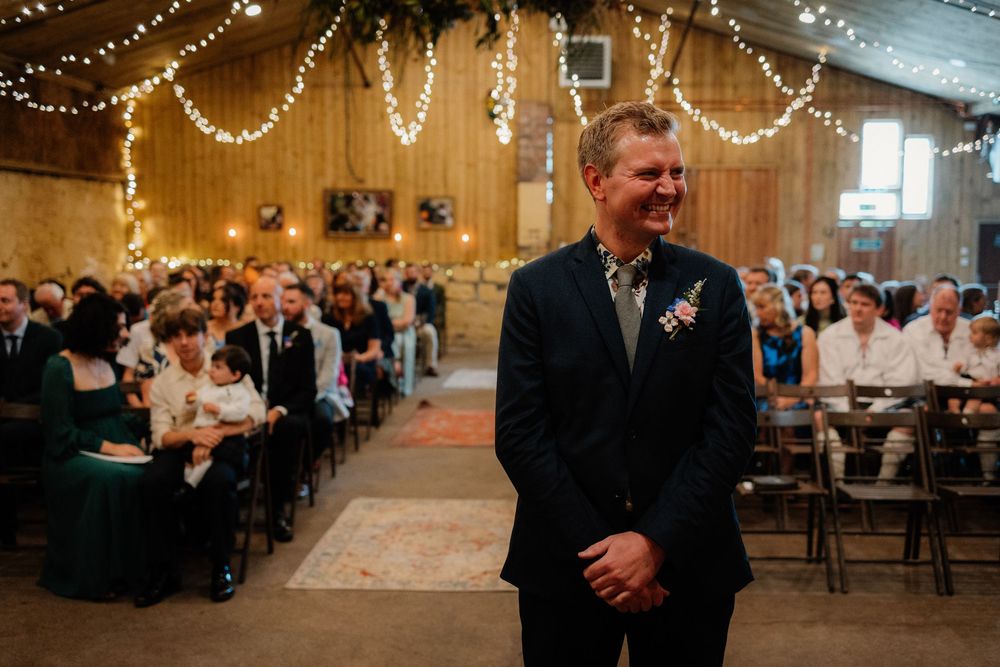 Groom smiling at wedding altar in barn venue with string lights and seated guests in background.