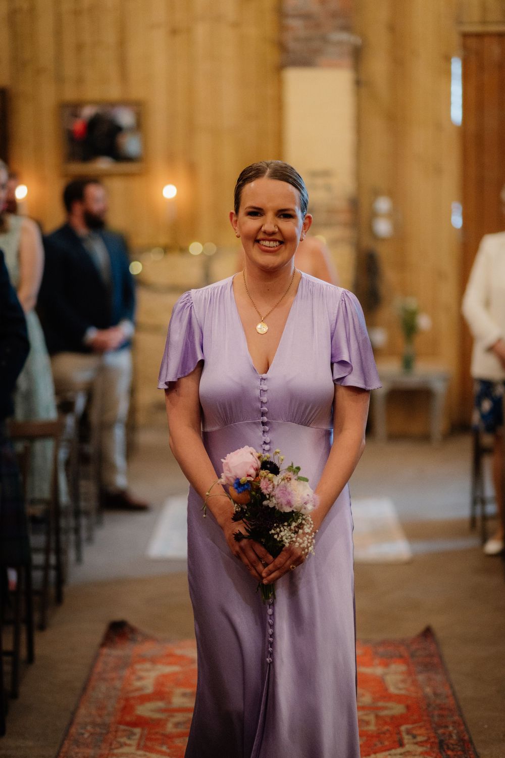 Smiling bridesmaid in lavender dress holding pink bouquet walking down aisle in rustic barn wedding venue.