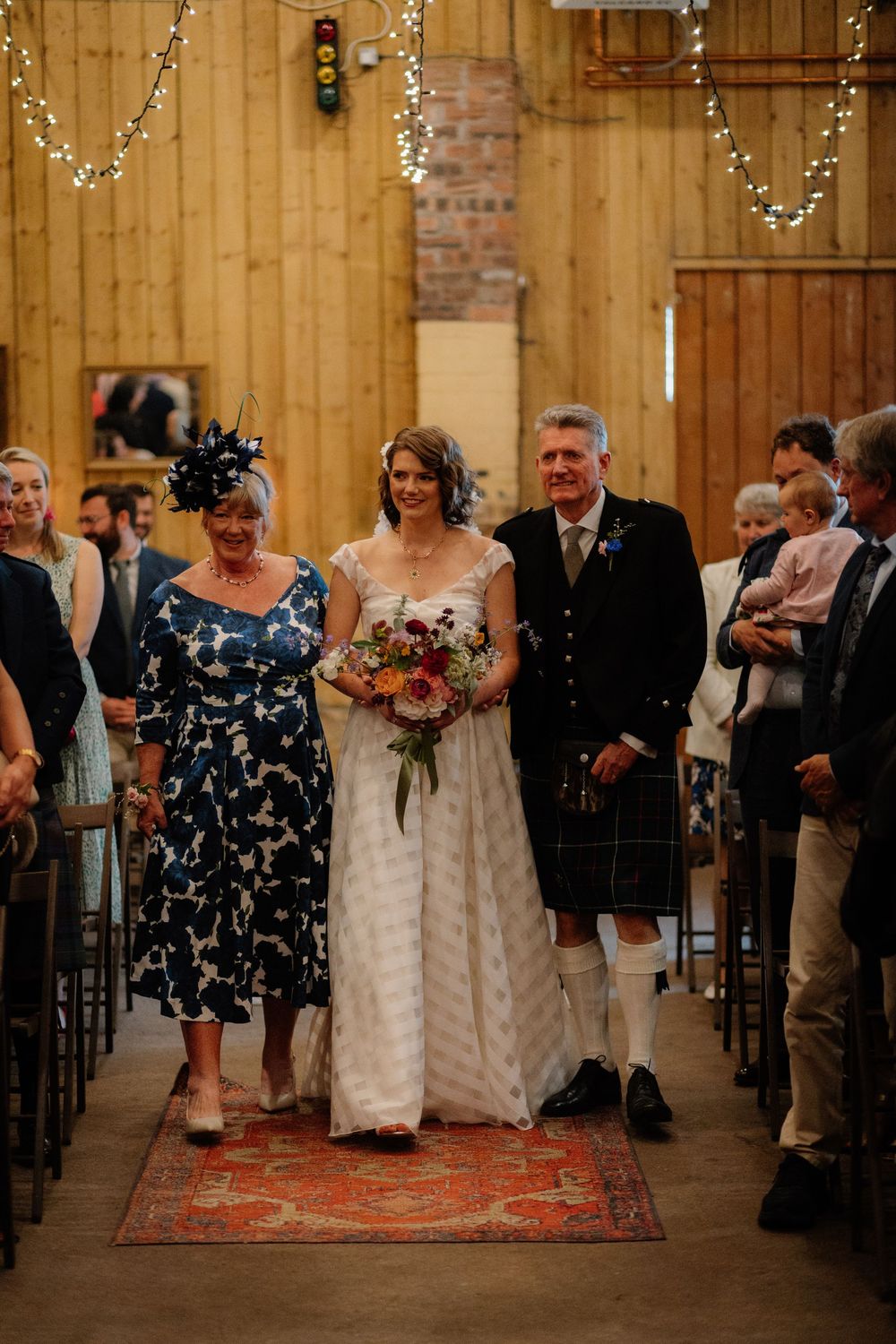 Bride walks down barn wedding aisle escorted by parents, wearing white gown with bouquet, guests seated on either side.