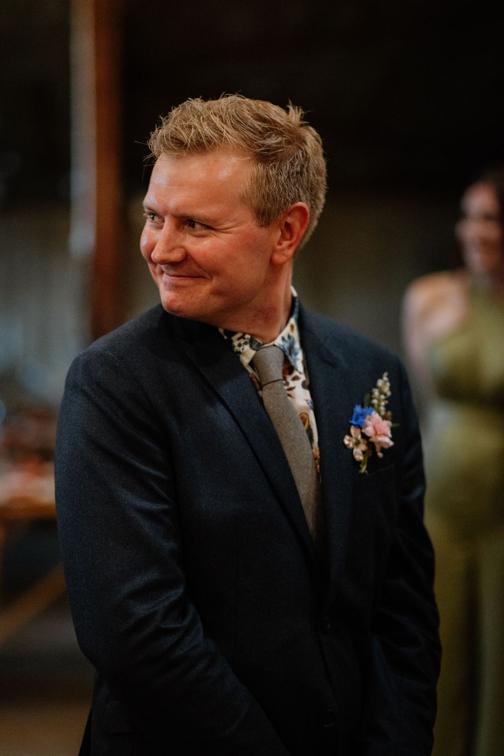 Smiling groom in dark suit with floral boutonniere and patterned tie at wedding ceremony.
