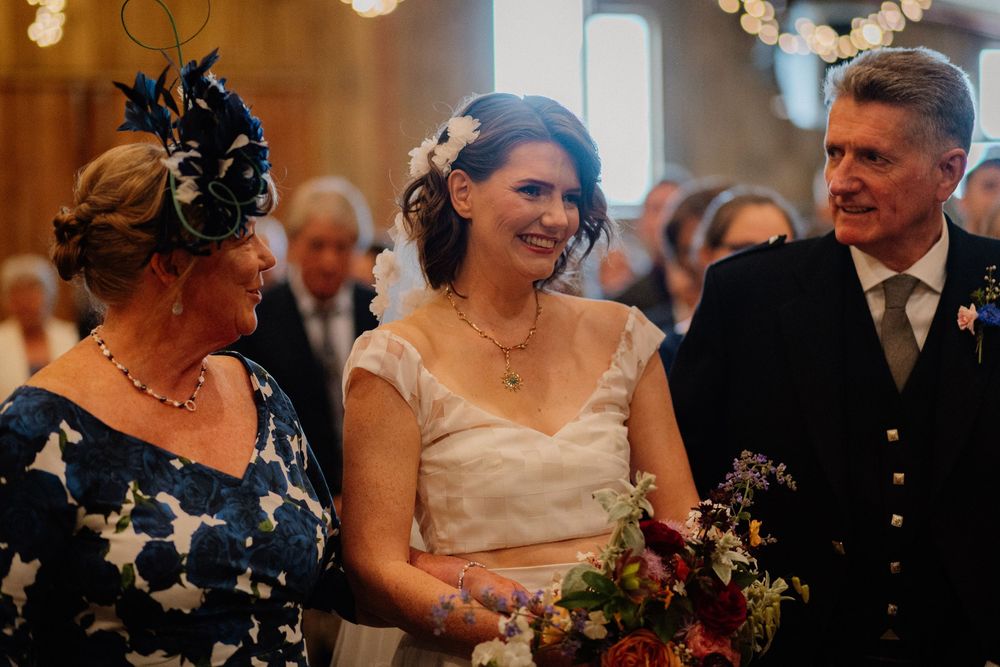 Bride smiling with bouquet flanked by family members in elegant wedding attire inside a bright venue.