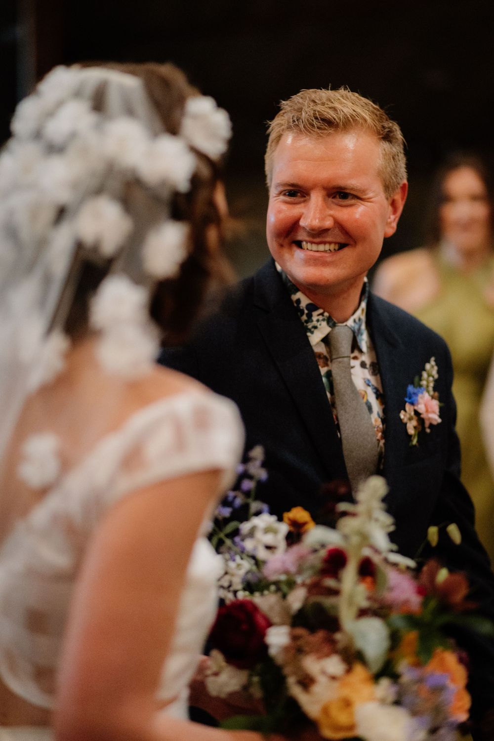 Smiling groom in navy suit with floral boutonniere faces bride in lace dress and floral veil at wedding ceremony.