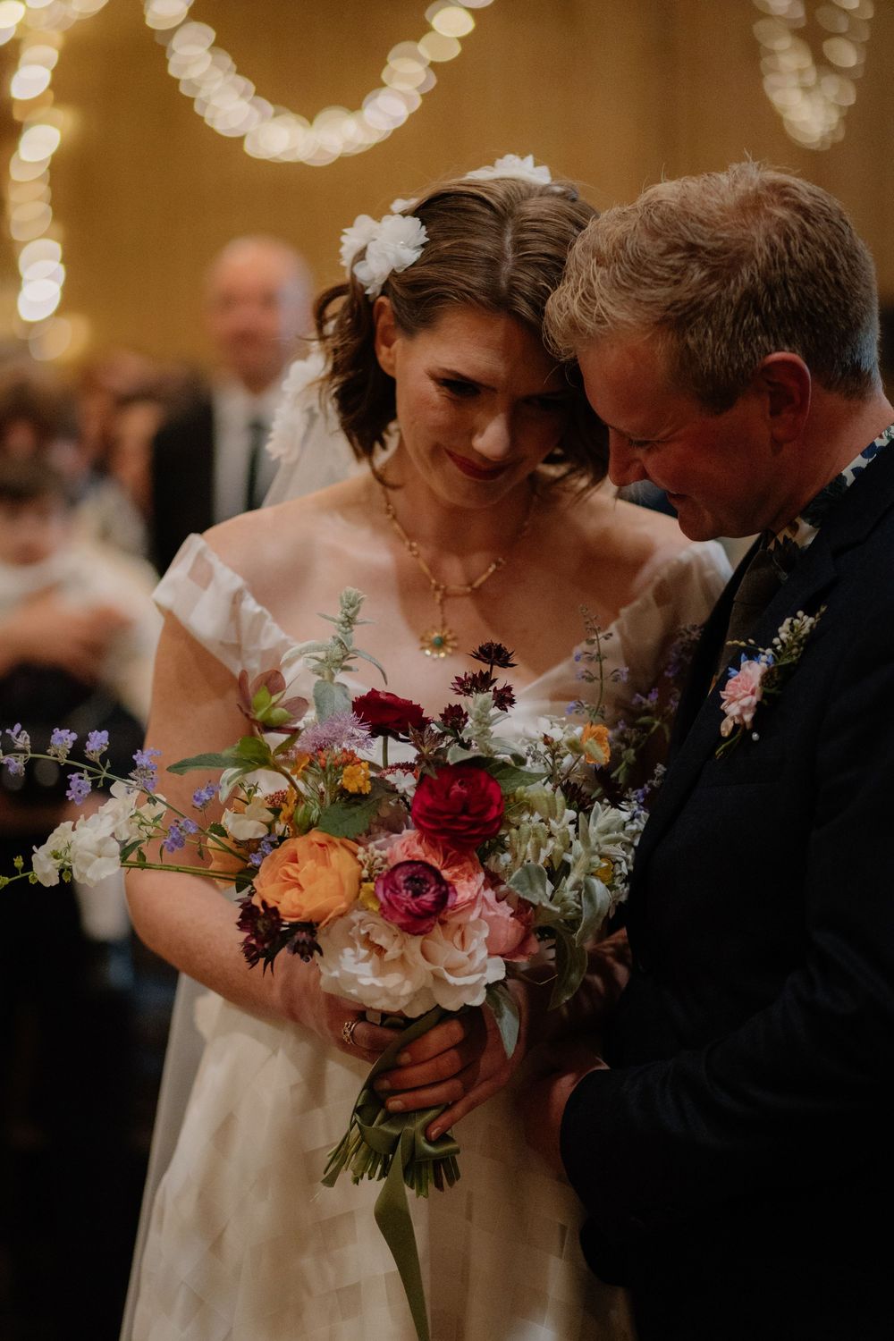 Bride holding colorful bouquet of ranunculus and wildflowers stands with older man in suit at warm wedding ceremony.