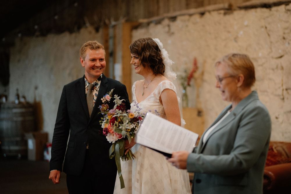 Smiling bride holding colorful bouquet stands with groom in navy suit during rustic barn wedding ceremony with officiant.
