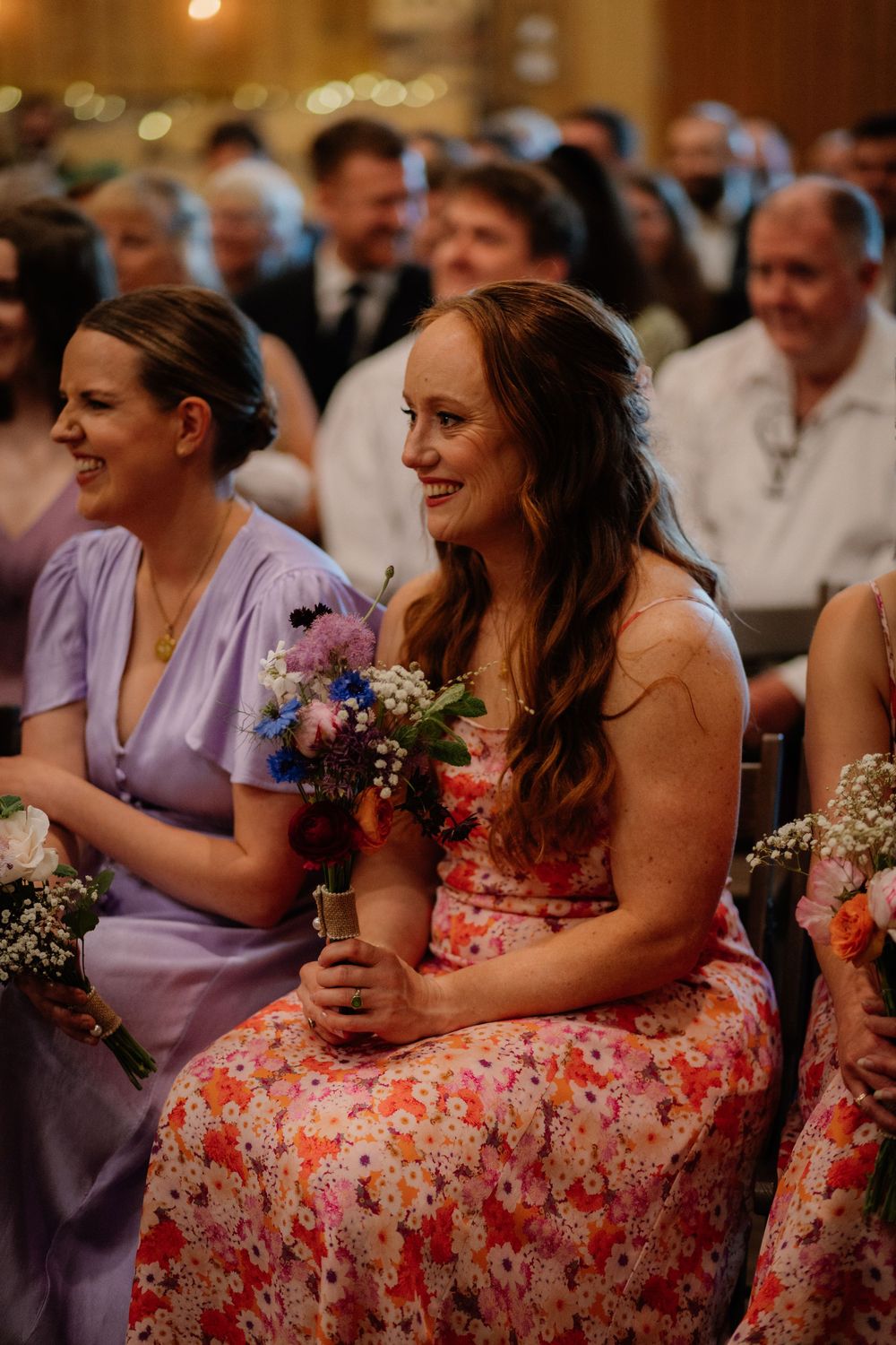 Smiling bridesmaid in floral dress holds colorful bouquet while seated at wedding ceremony.