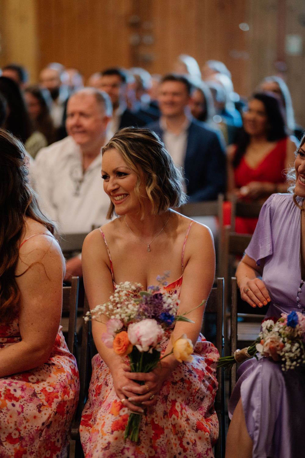Smiling bridesmaid in floral dress holds colorful bouquet while seated at a wedding ceremony indoors.