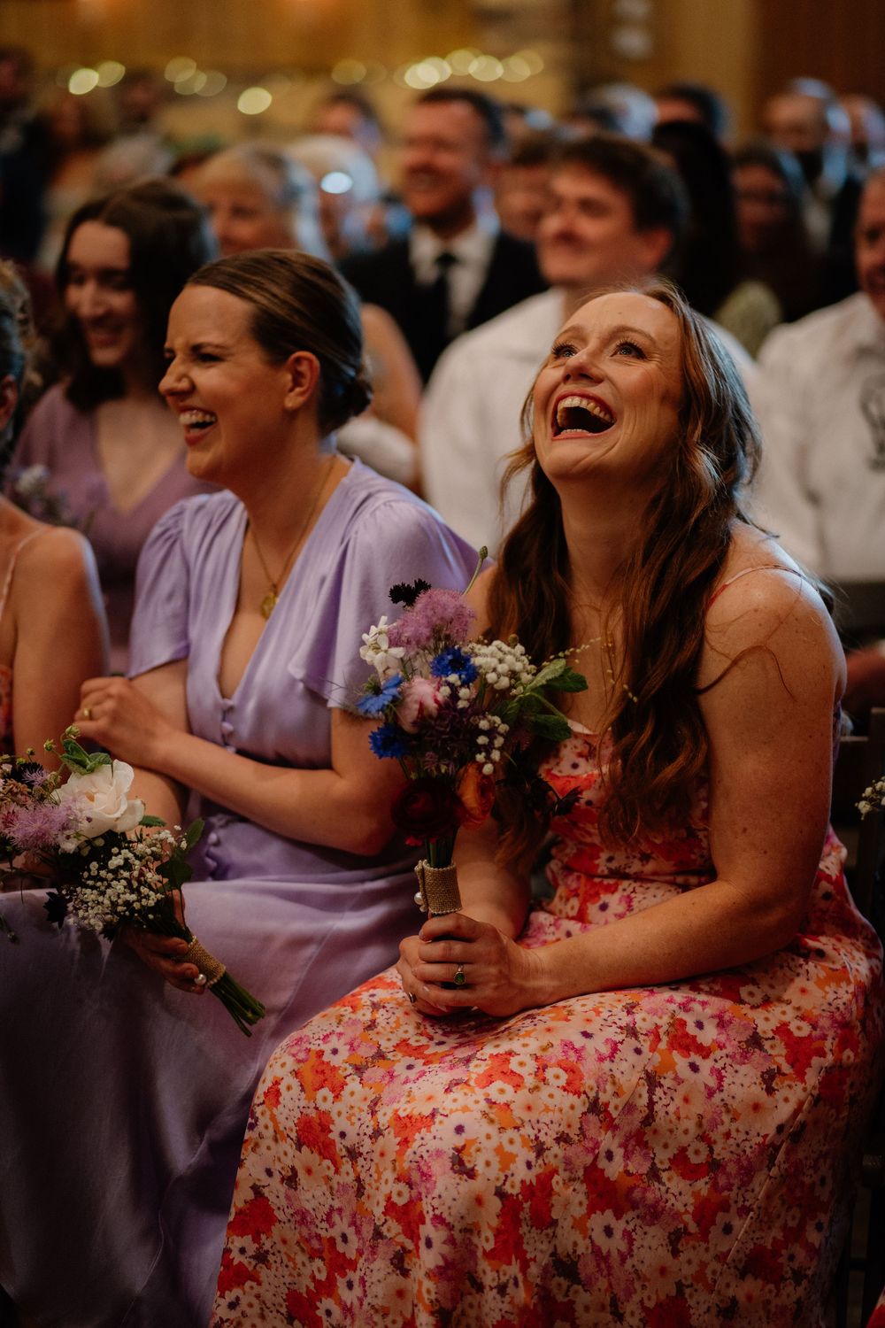 Bridesmaids laughing joyfully during wedding ceremony, wearing purple and floral dresses while holding colorful bouquets.