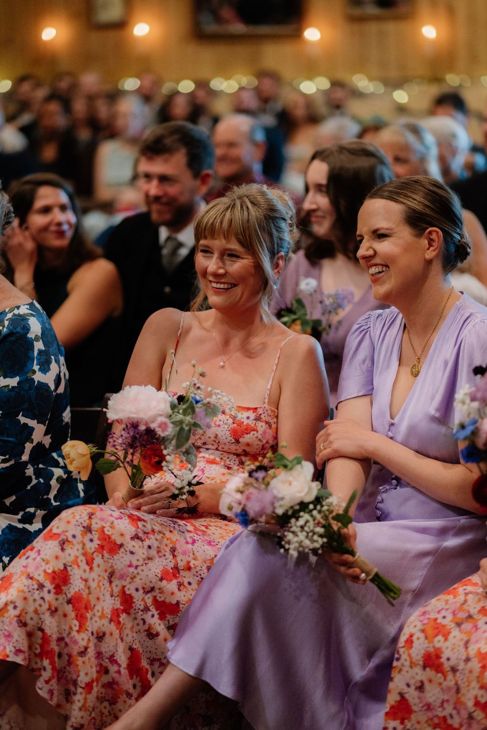Two bridesmaids in floral and lavender dresses holding bouquets, smiling during a wedding ceremony.