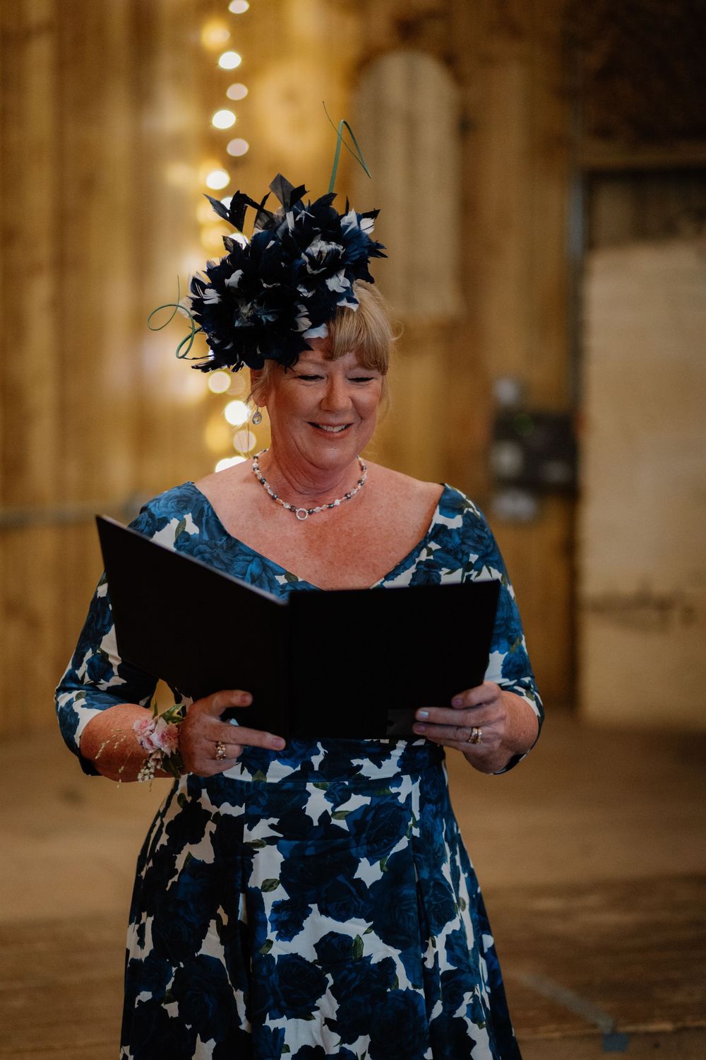 Woman in blue floral dress and black fascinator hat smiling while reading from a folder at a wedding ceremony.