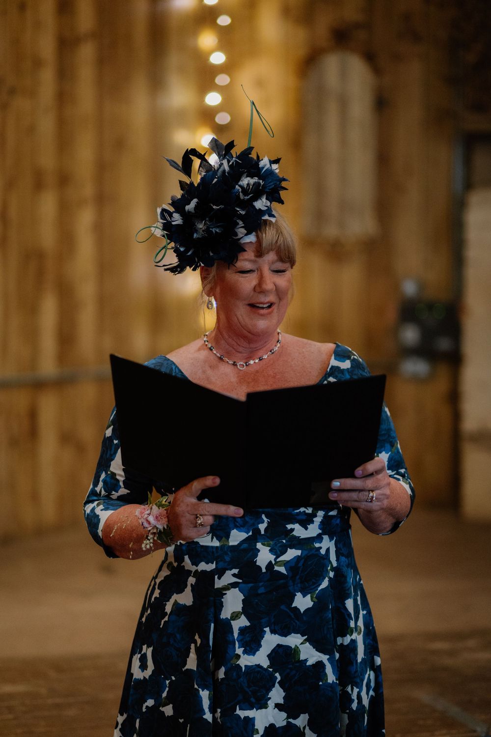 Woman in blue floral dress and black fascinator hat reads from a black folder at a formal event.