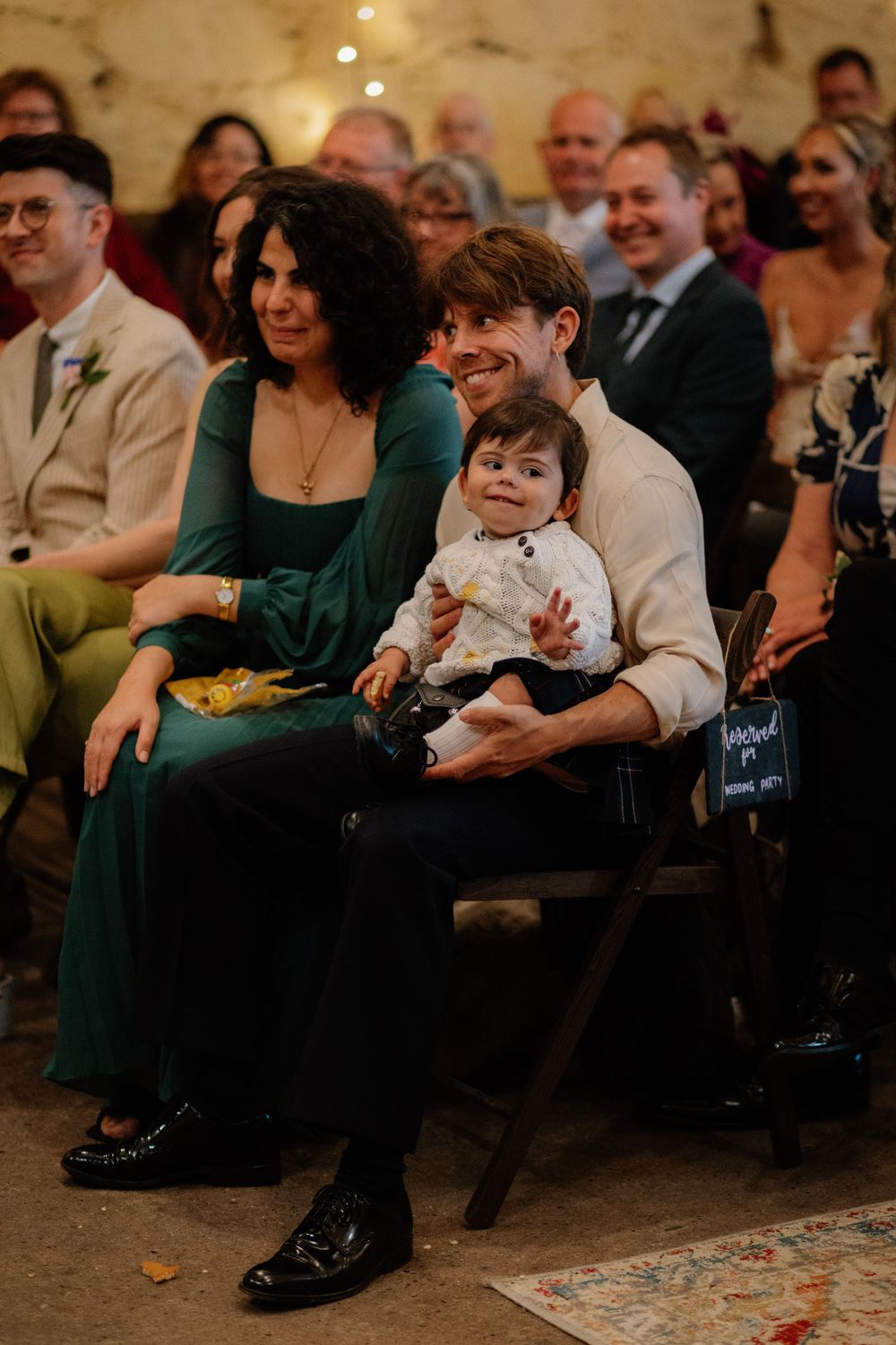 Man holds baby at wedding ceremony beside woman in green dress, all smiling warmly.