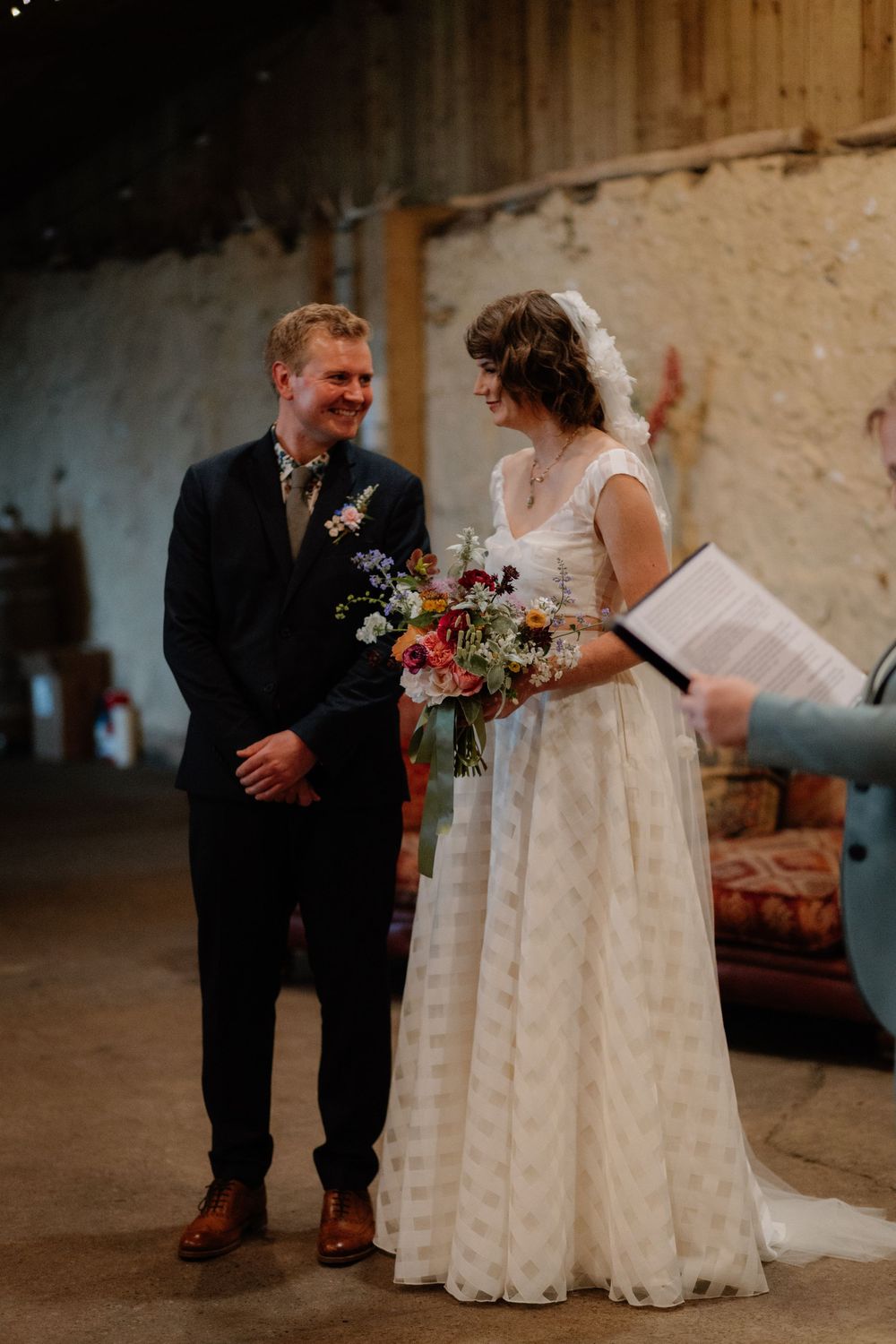 Bride and groom smiling during barn wedding ceremony, bride holding vibrant wildflower bouquet in elegant white gown.