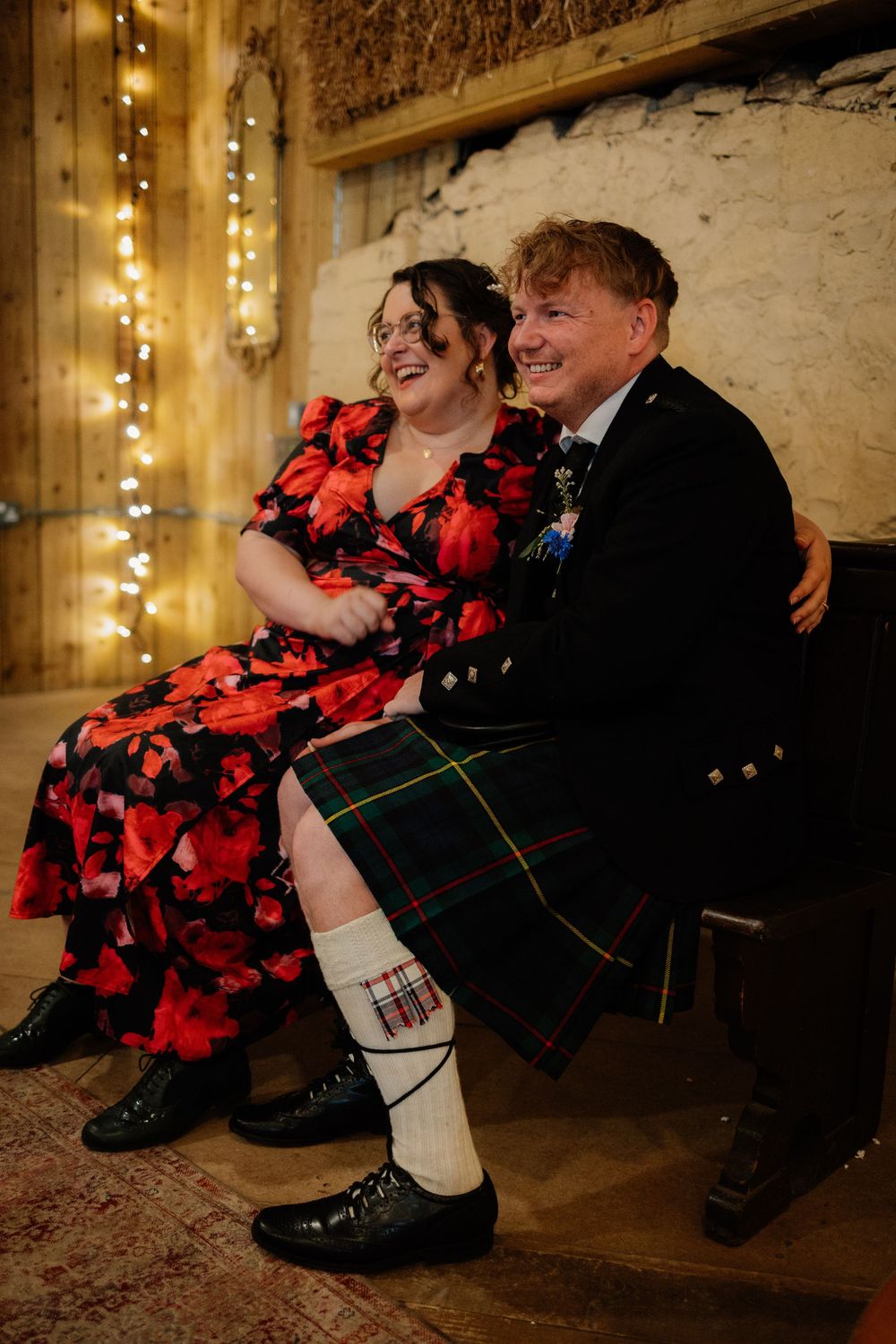 Couple laughing together at wedding, woman in red floral dress, man in traditional Scottish kilt and black jacket.