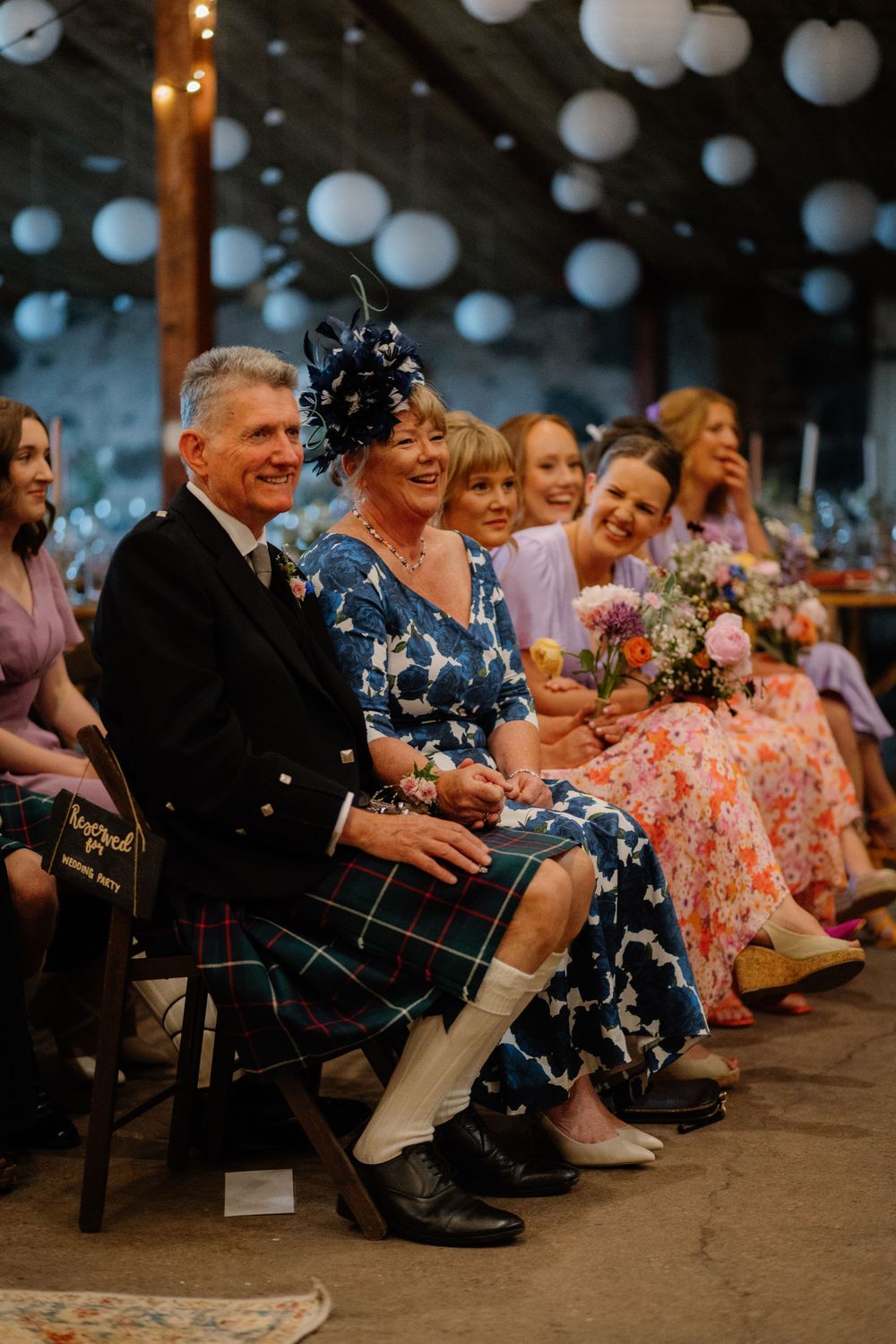 Wedding guests seated in barn venue, man in tartan kilt and woman in blue floral dress with fascinator smiling.