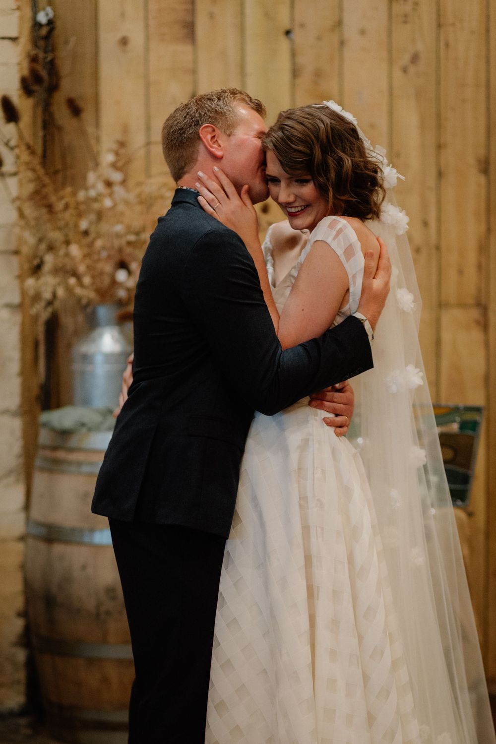 A bride and groom share a romantic embrace in a rustic barn setting, with the bride in a white gown and veil.