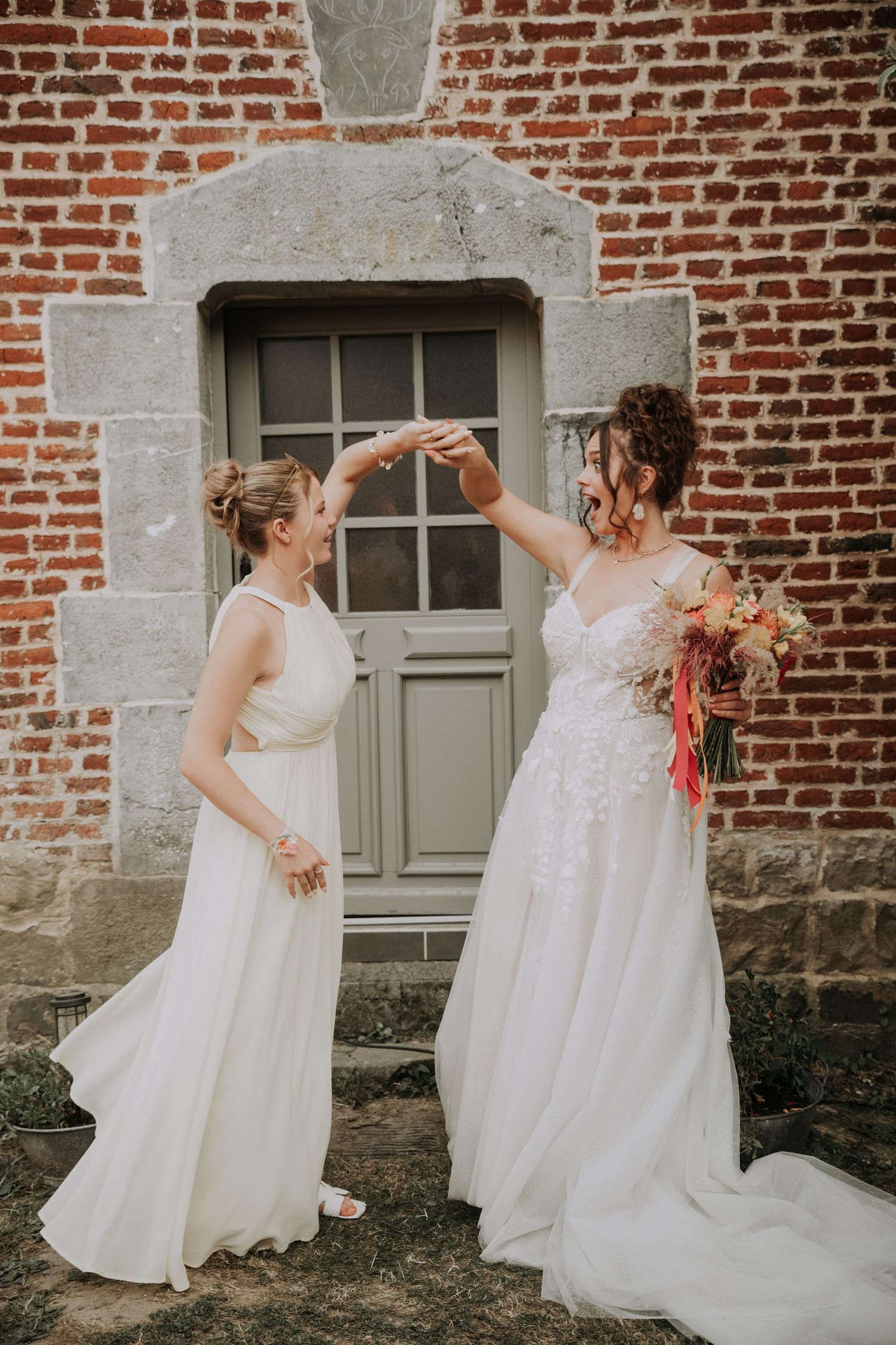 Two brides in white wedding gowns dancing together outside a brick building with a gray door.