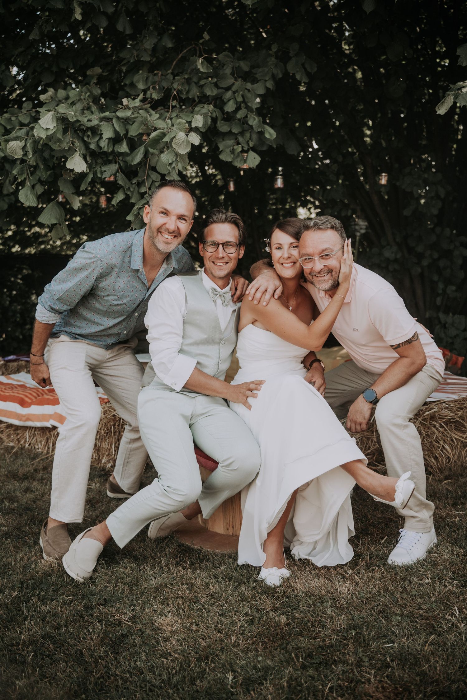 Four friends pose joyfully at an outdoor wedding, sitting on hay bales under trees in summer attire.