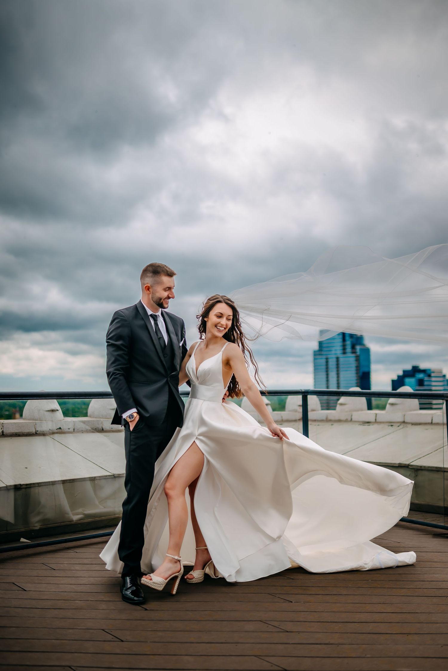 Bride and groom dancing on rooftop terrace with flowing wedding dress and veil against dramatic cloudy city skyline.