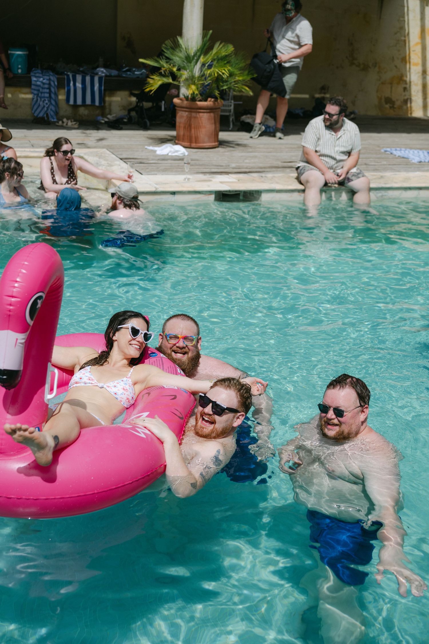 Group of friends enjoying a sunny pool party with a pink flamingo float in a resort swimming pool.