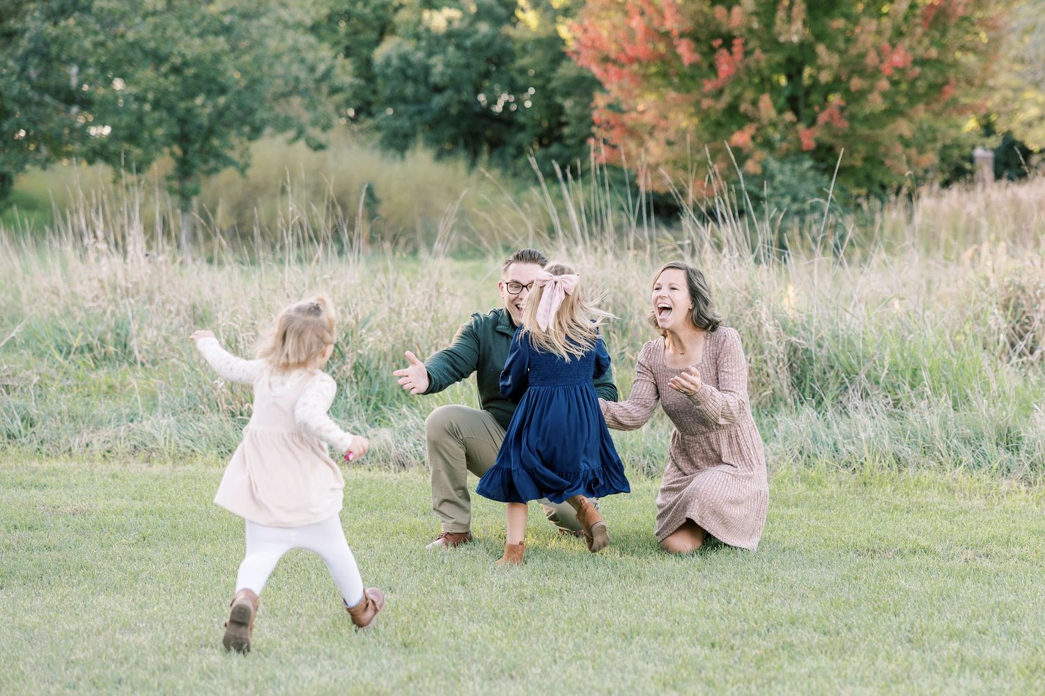 Family of four playing joyfully outdoors on green grass with colorful autumn trees in background.