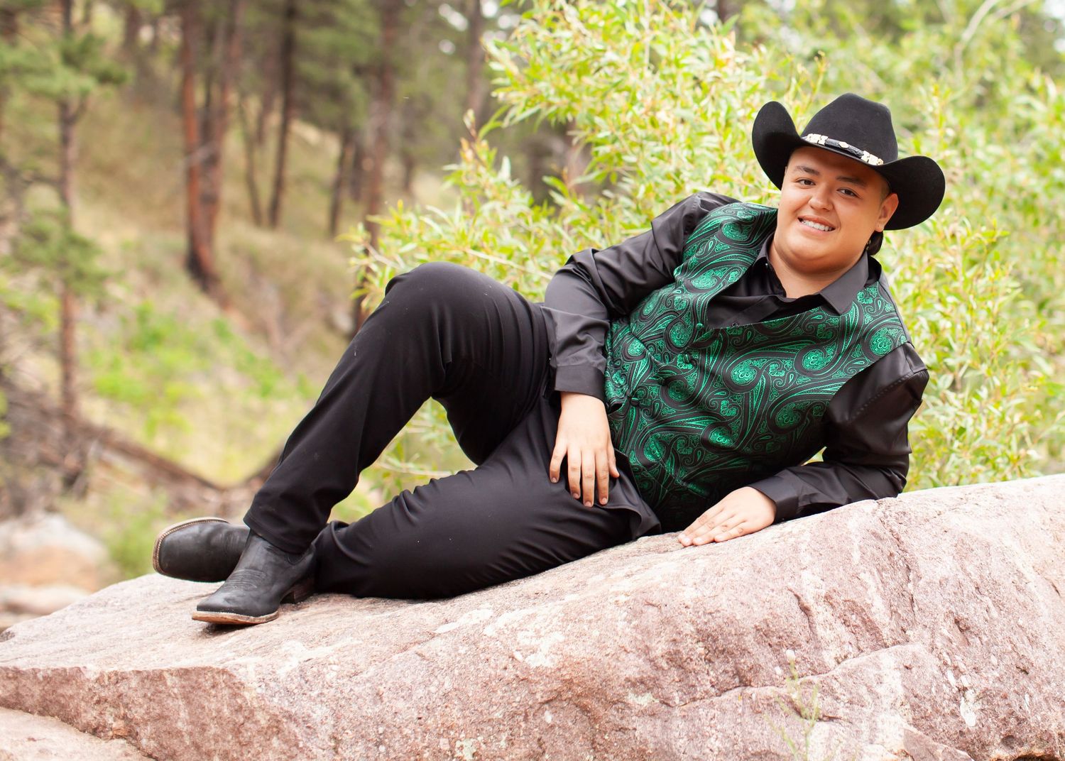 Person in black cowboy hat and green floral shirt relaxing on rocks in a scenic mountain forest setting.