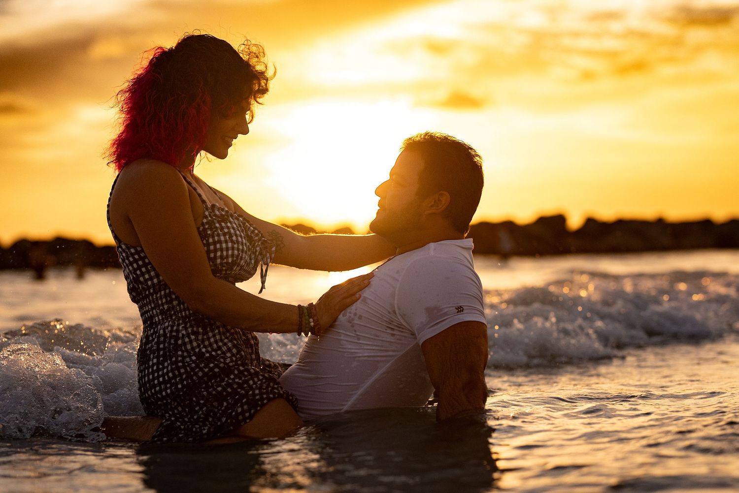 Couple sitting together in shallow ocean waves silhouetted against a vibrant golden sunset.
