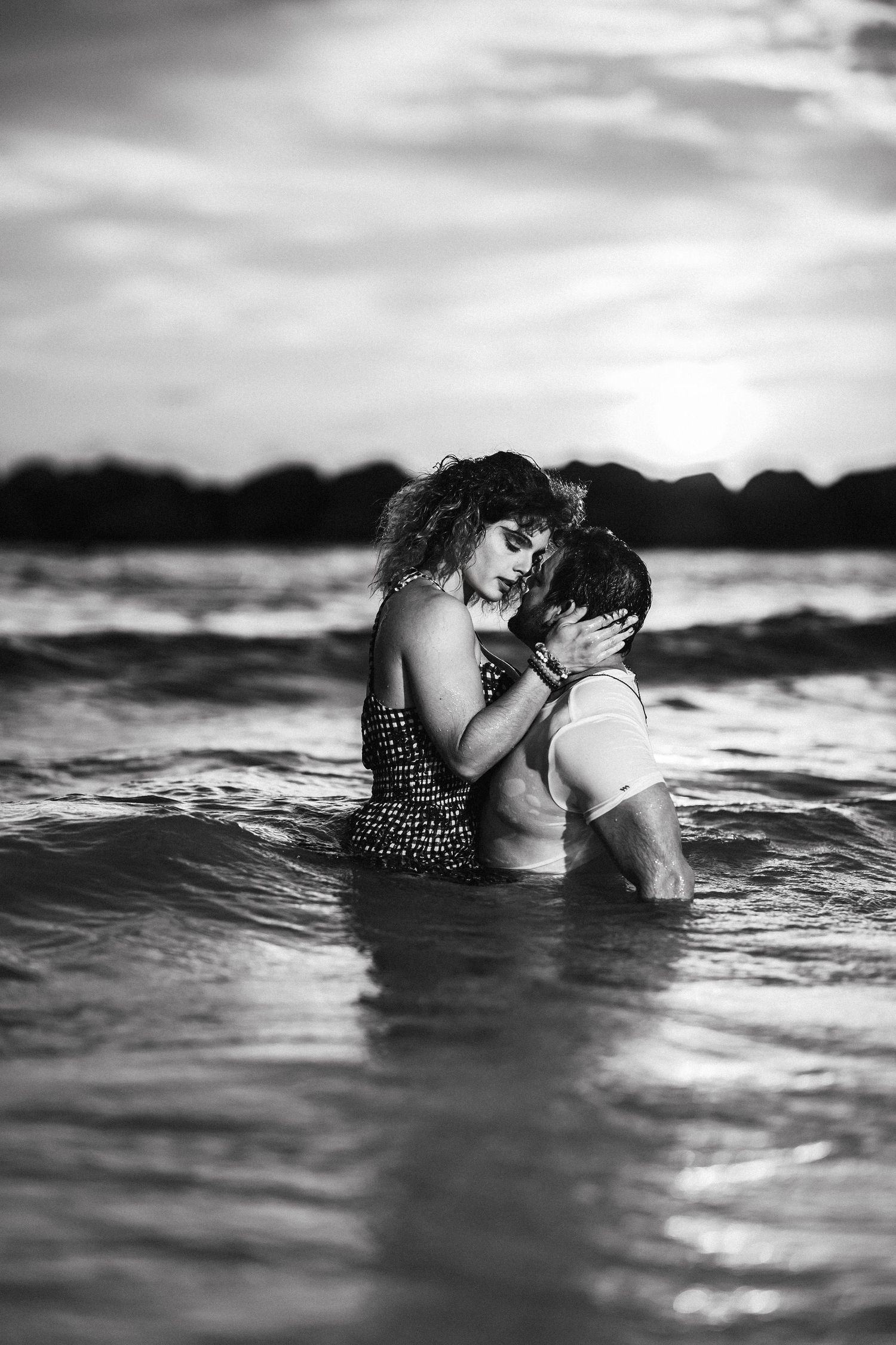 Romantic black and white photo of couple embracing in ocean waves at sunset.