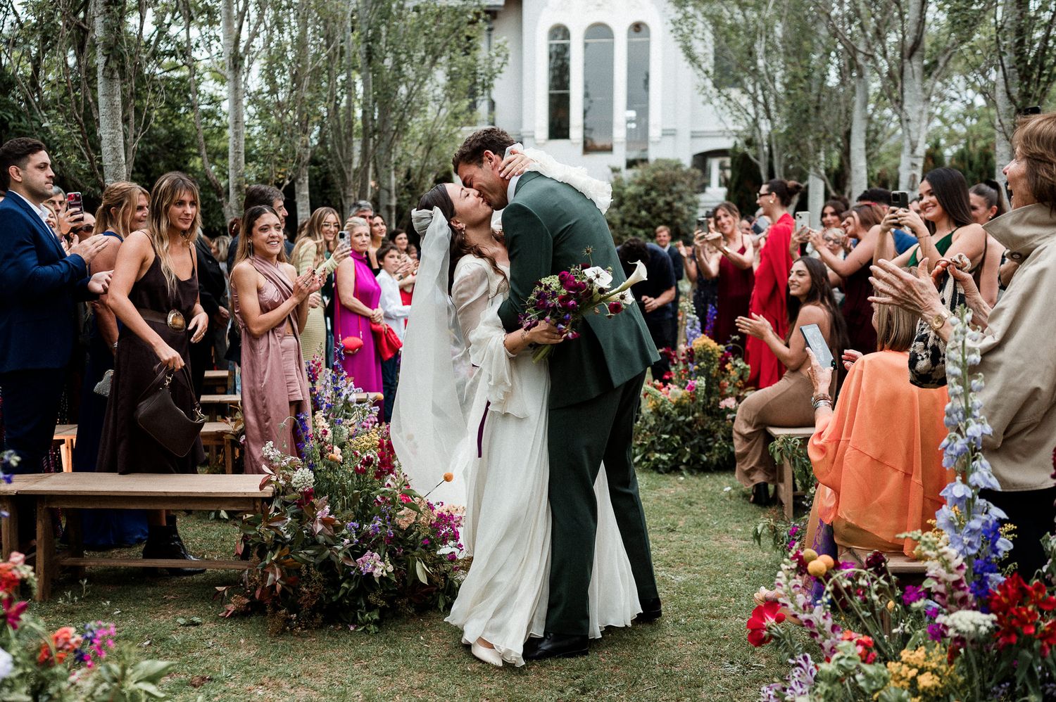 Outdoor wedding ceremony with guests celebrating a couple's first kiss under trees near a white church.