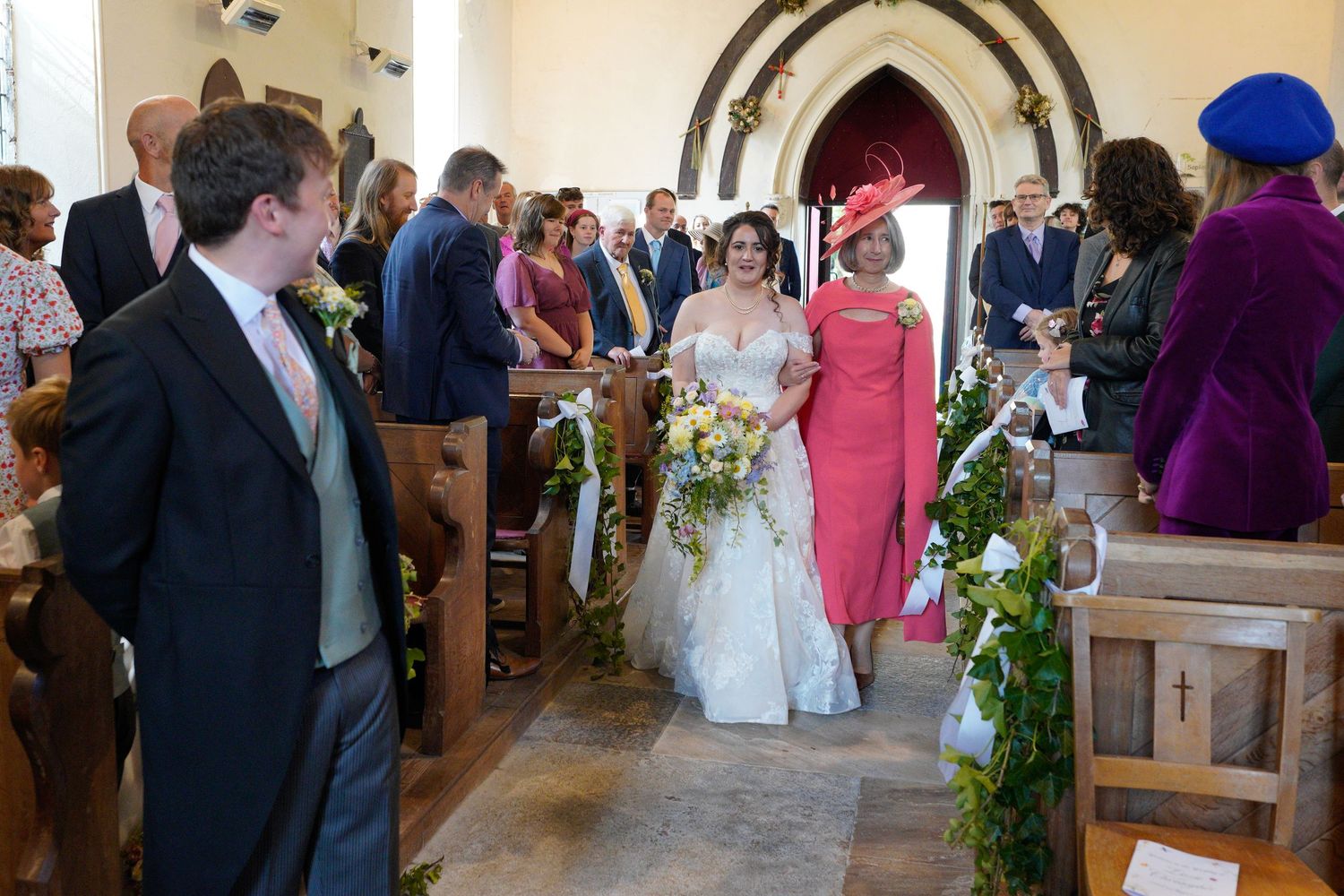 Bride walks down church aisle in lace gown, escorted by woman in pink dress, with guests seated in wooden pews.