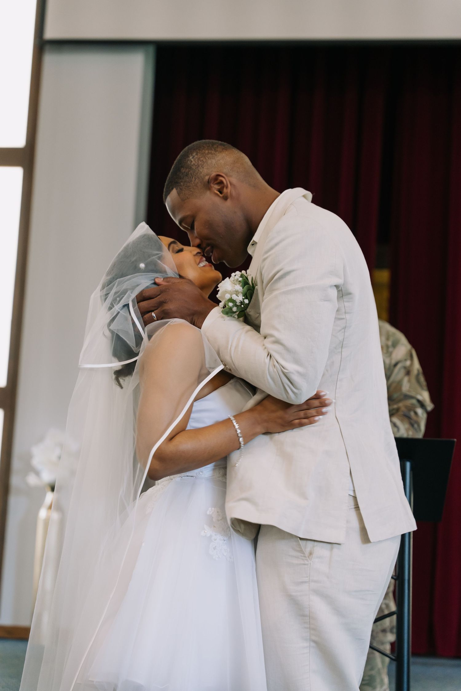 A Black bride and groom share their first kiss at the altar, bride in white gown with veil, groom in beige suit with boutonniere.