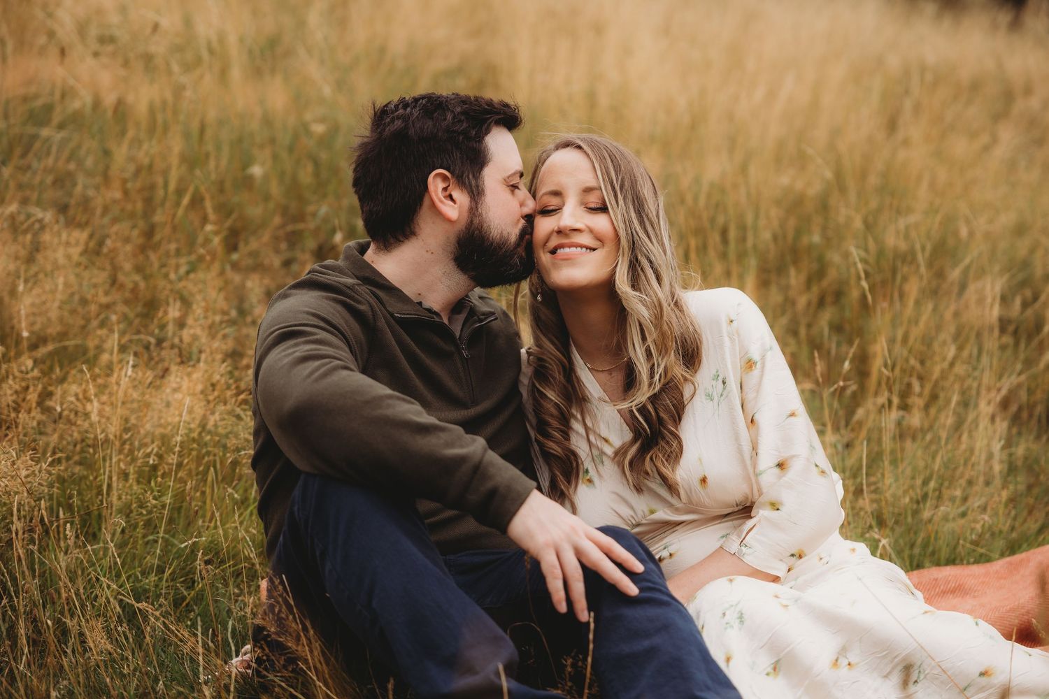 Couple embracing outdoors, pregnant woman in floral dress, man kissing her cheek in a lush garden setting.